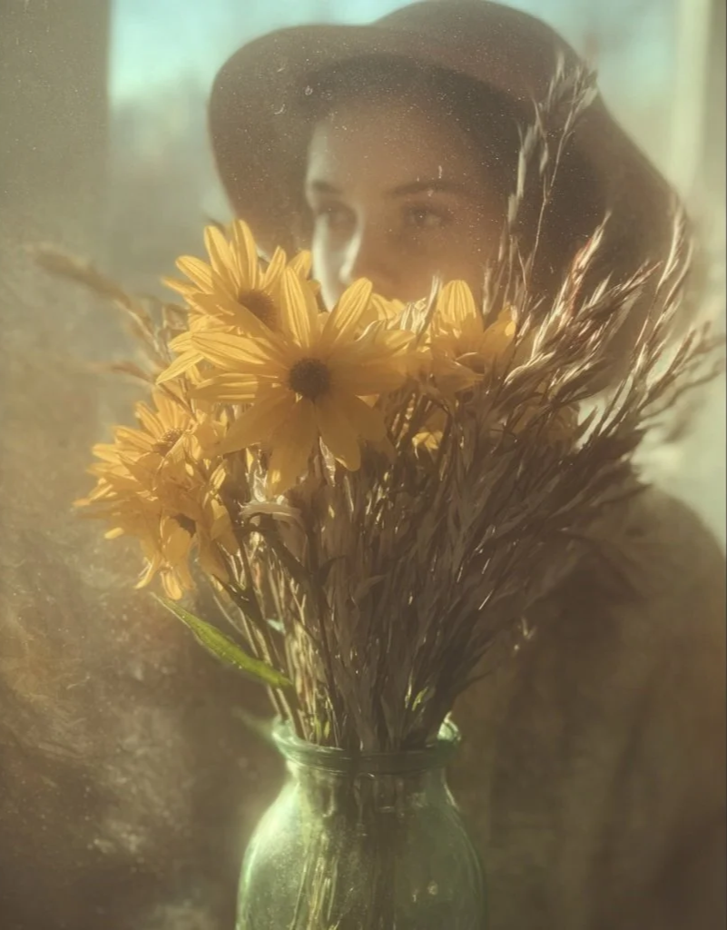A woman in a hat holding a bouquet of yellow flowers in a glass vase.