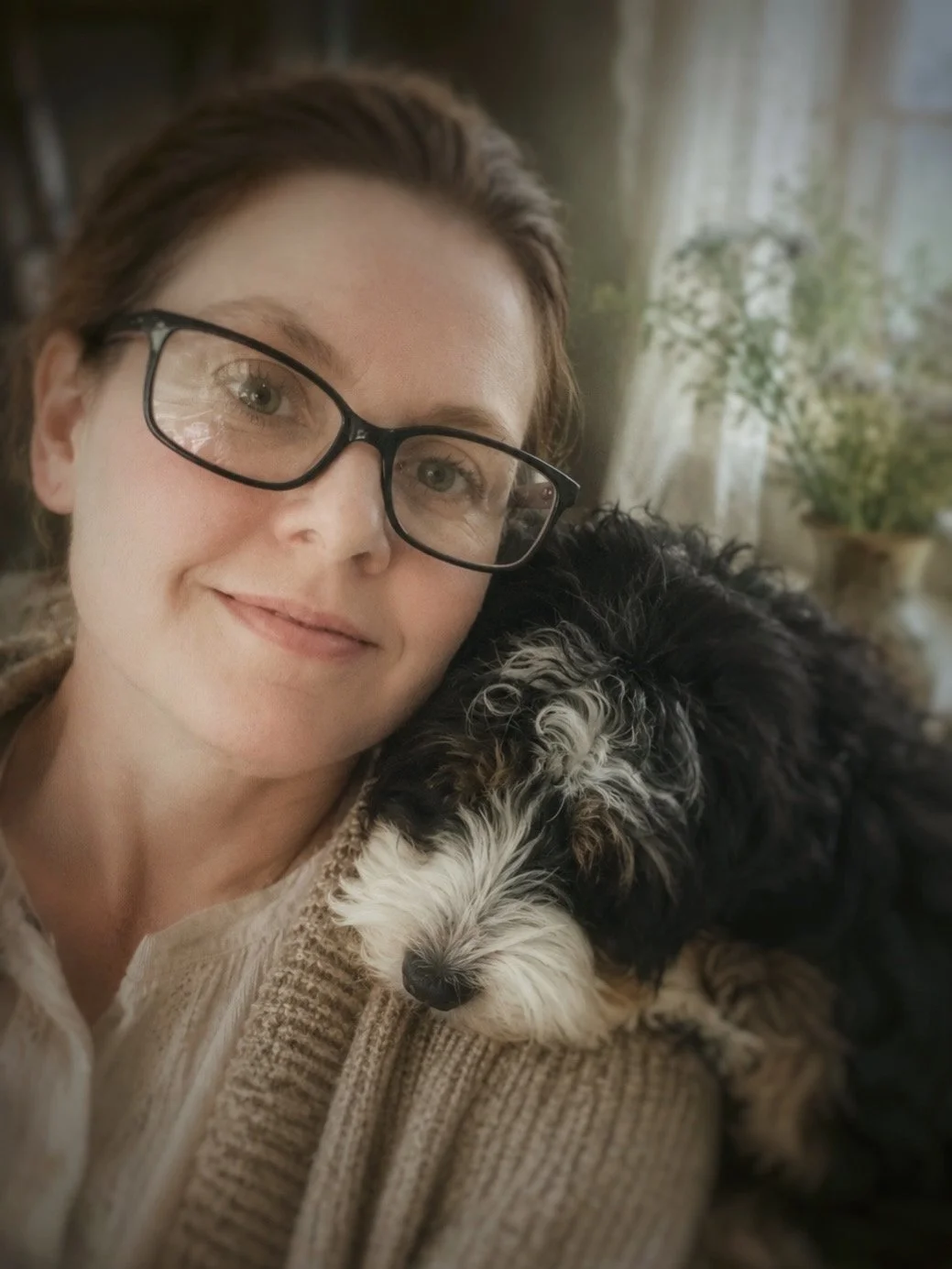 A woman with glasses smiling next to a black and white curly-haired dog resting its head on her shoulder indoors, with a window and houseplants in the background.