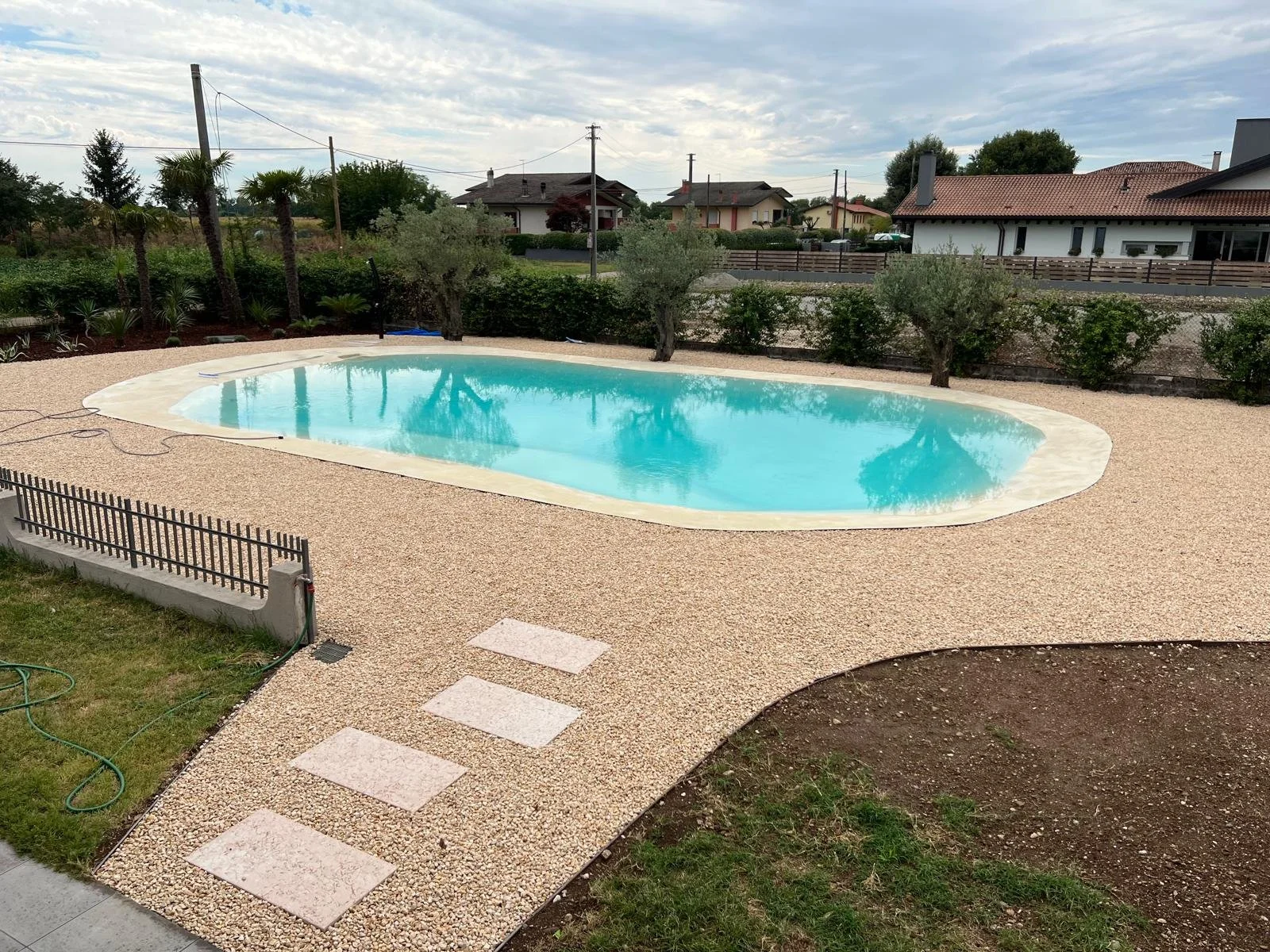 Piscina rotonda in giardino con una pavimentazione di ghiaia, alberi e case sullo sfondo, cielo nuvoloso.