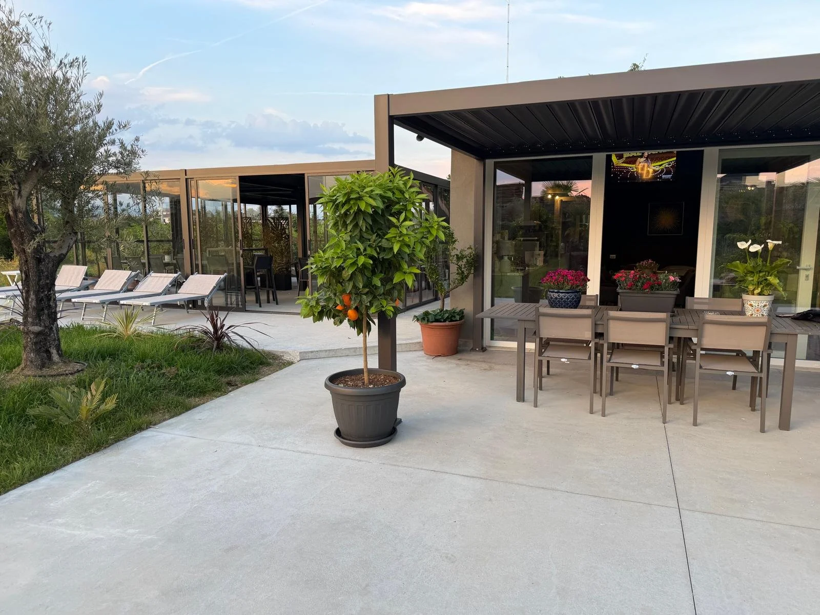 Balcone con tavolo e sedie, piante in vaso, area relax con ombrelloni e sdraio, in un giardino con alberi e prato.