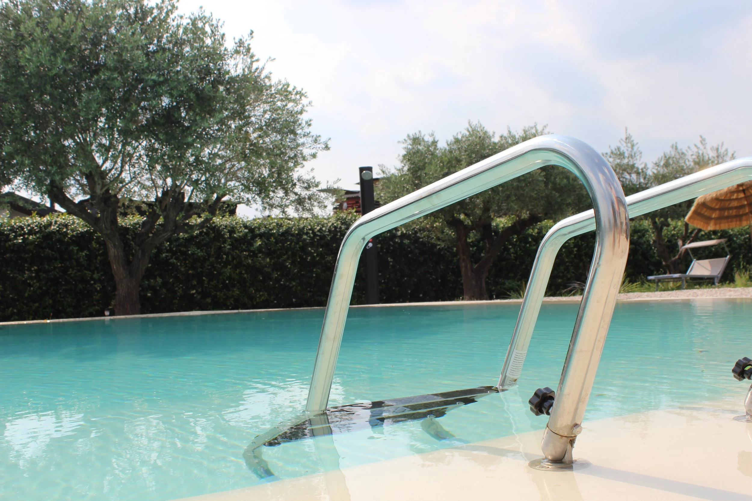 Piscina con scaletta in metallo, acqua blu e intorno alberi e cielo con nuvole leggere.