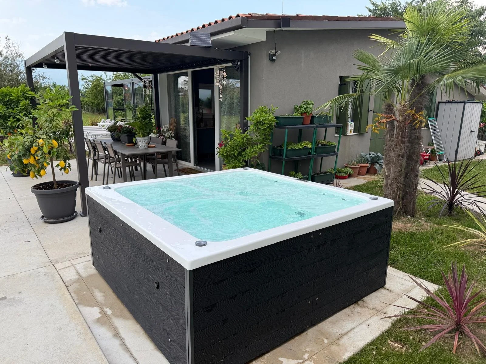 Una terrazza con una vasca idromassaggio, un tavolo per mangiare all'aperto con sedie, piante in vaso e un'area coperta, vicino a un muro con una finestra e alcune piante tropicali.