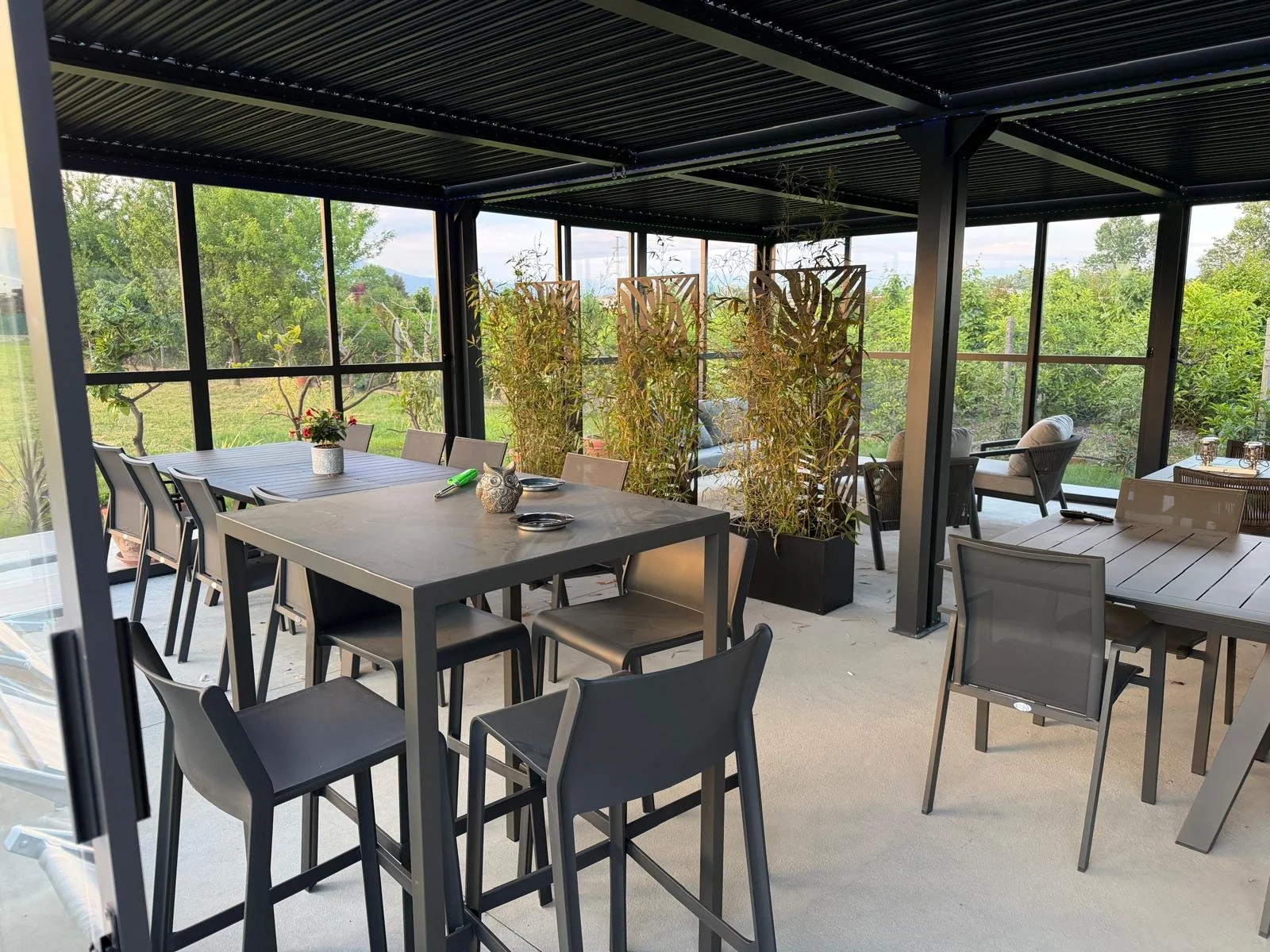 Veranda con tavoli e sedie, piante alte in vasi, finestre panoramiche che mostrano un giardino verde con alberi e un cielo nuvoloso.