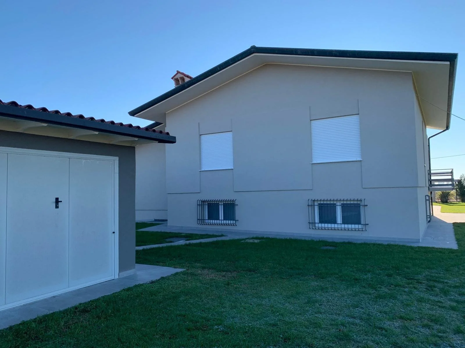 Casa moderna con finestre chiuse da tapparelle bianche, recinzioni di sicurezza alle finestre inferiori, e un giardino di prato verde, sotto un cielo chiaro.