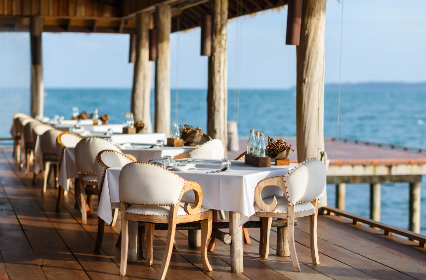 Dining table setup on a wooden deck overlooking the ocean, with chairs and tableware.