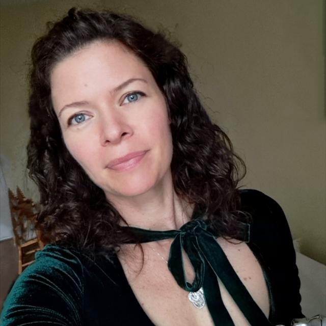 Eine junge Frau mit lockigem braunem Haar, blauen Augen und leichtem Make-up trägt ein dunkelgrünes Kleid und eine silberne Halskette, posiert in einem Raum mit gelber Wand im Hintergrund.