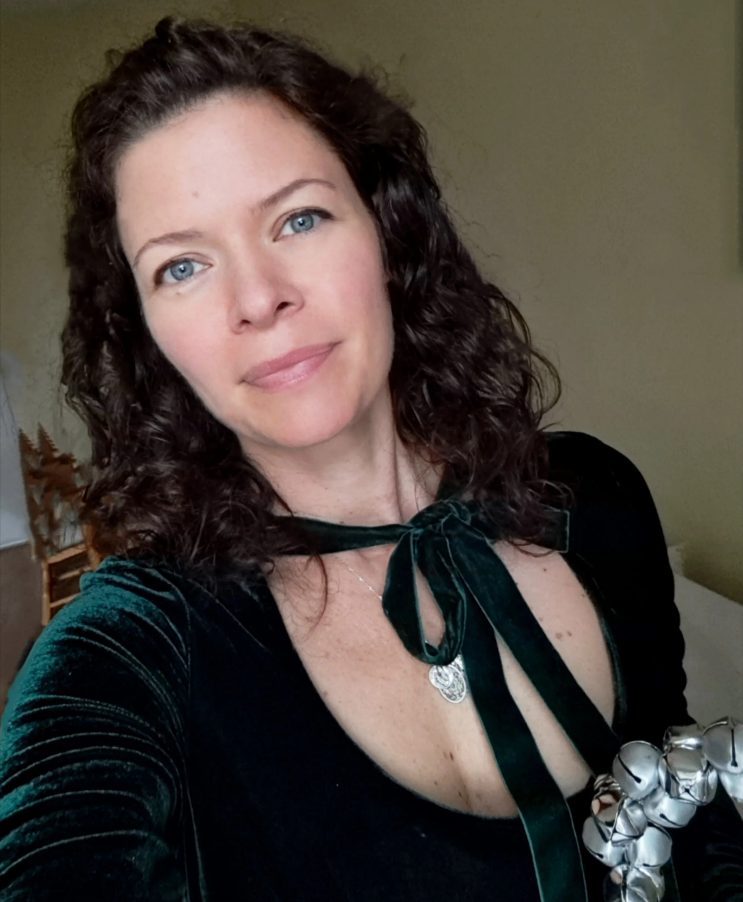Eine junge Frau mit lockigem braunem Haar, blauen Augen und leichtem Make-up trägt ein dunkelgrünes Kleid und eine silberne Halskette, posiert in einem Raum mit gelber Wand im Hintergrund.