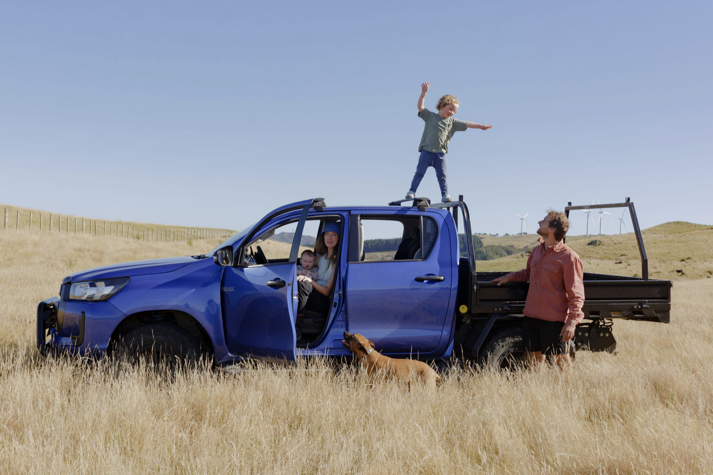 A family with two children and a dog in a rural open field next to a blue pickup truck with its doors open. One child is standing on the roof of the truck with arms raised, the mother is sitting inside the truck holding a baby, and the father is standing next to the truck. Wind turbines are visible in the background under a clear blue sky.