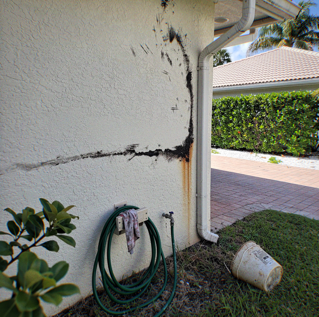 Scorch marks on a stucco wall above a coiled hose and faucet, with an overturned bucket on grass and a hedge nearby.