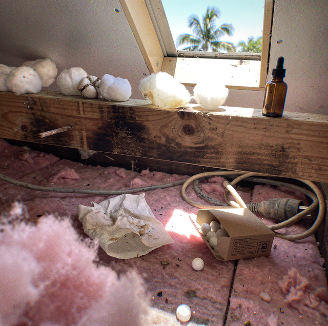 Cotton balls on a wooden beam by an open window, with insulation, a cord, a dropper bottle, and a box of white pills below.