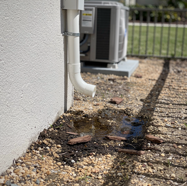 White wall with downspout drains water onto gravel beside HVAC unit and metal fence.