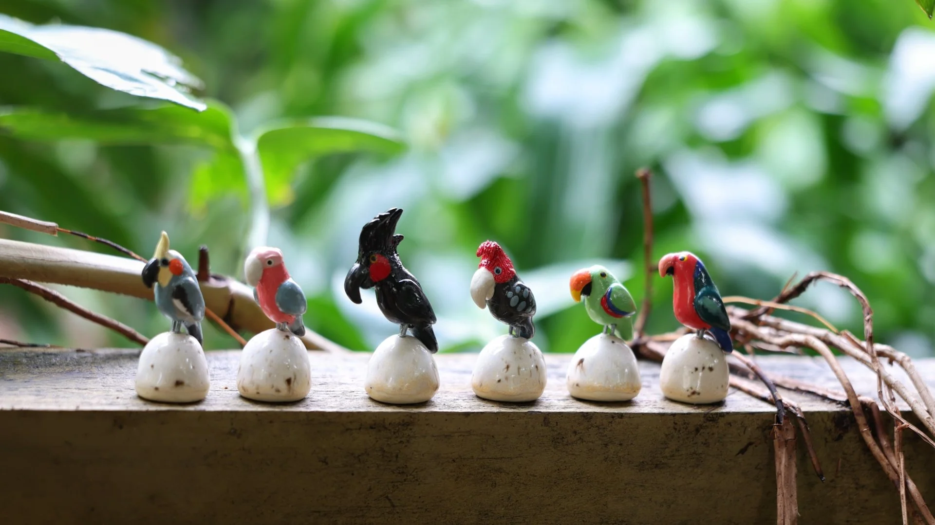 Six colorful small bird figurines with various designs, standing on white bases lined up on a wooden surface with green leaves in the background.