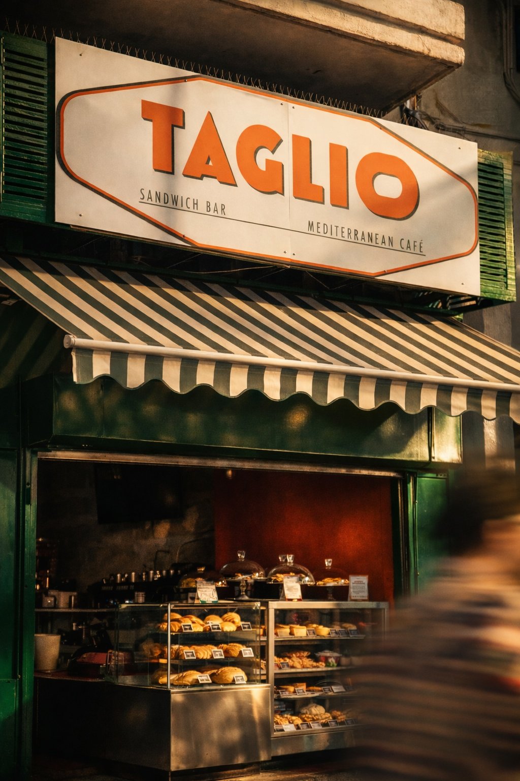 The storefront of Taglio, a sandwich bar and Mediterranean café, with a sign and striped awning, displaying baked goods inside.
