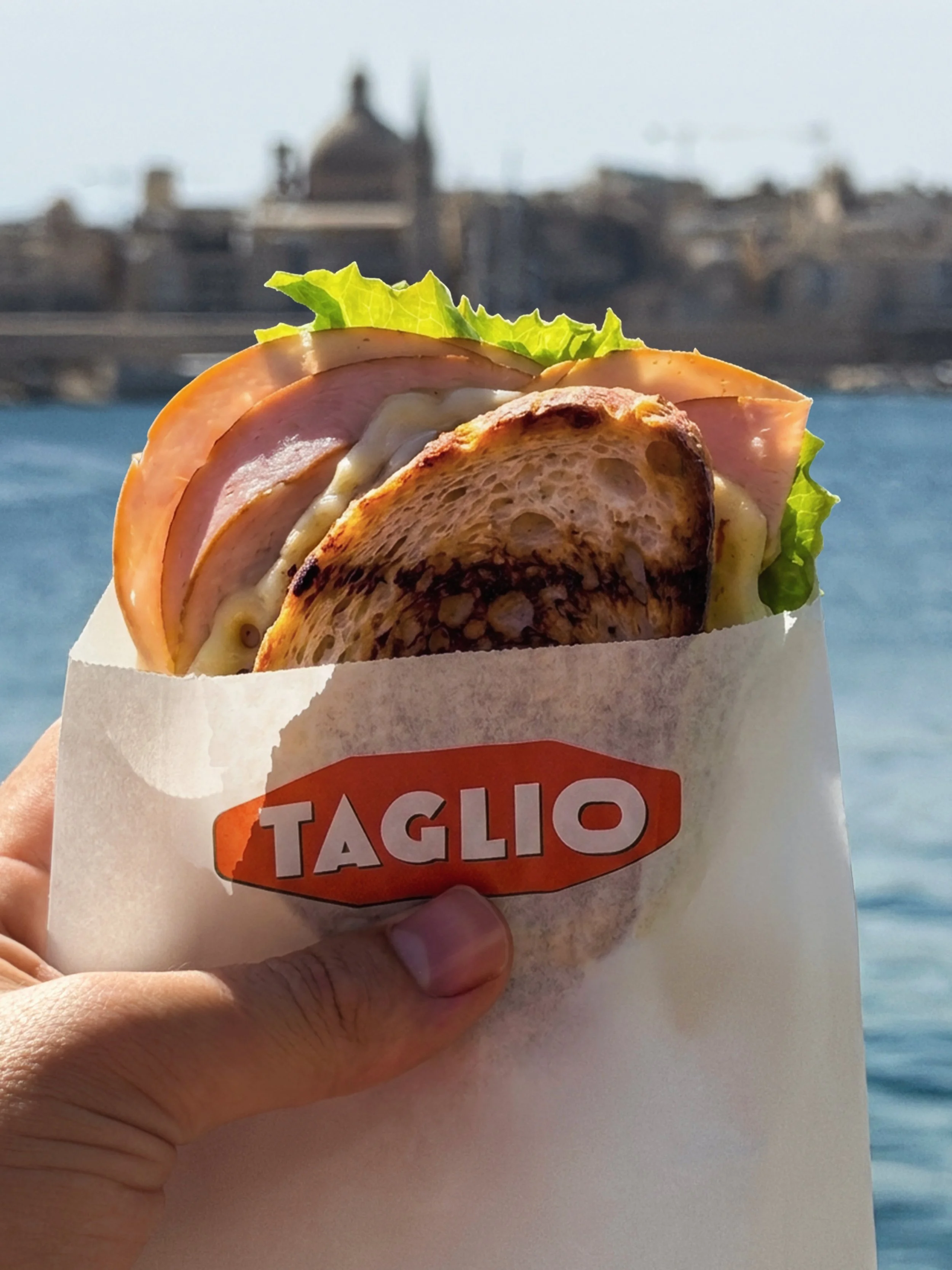 A sandwich with meat, cheese, lettuce, and a bun, held by a hand in front of a cityscape with water and historical buildings in the background.