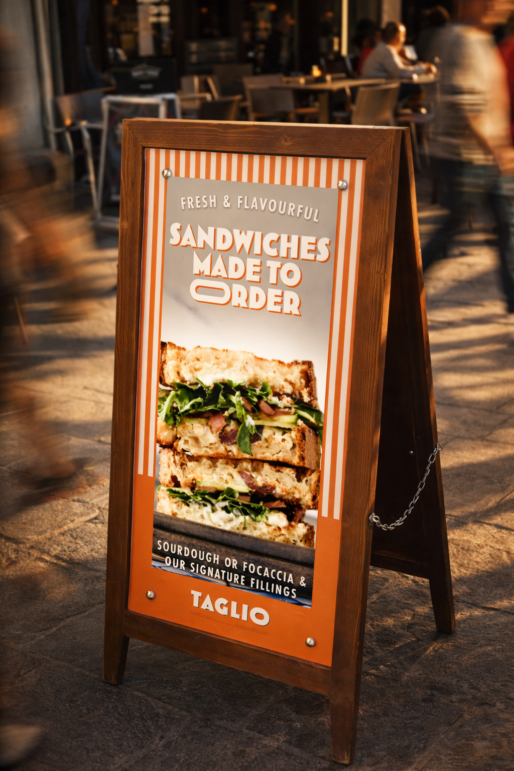 Outdoor restaurant sign advertising sandwiches made to order, featuring a large photo of a sandwich with greens and meats. The sign emphasizes fresh and flavorful ingredients, with options for sourdough, focaccia, and signature fillings, branded by Taglio.