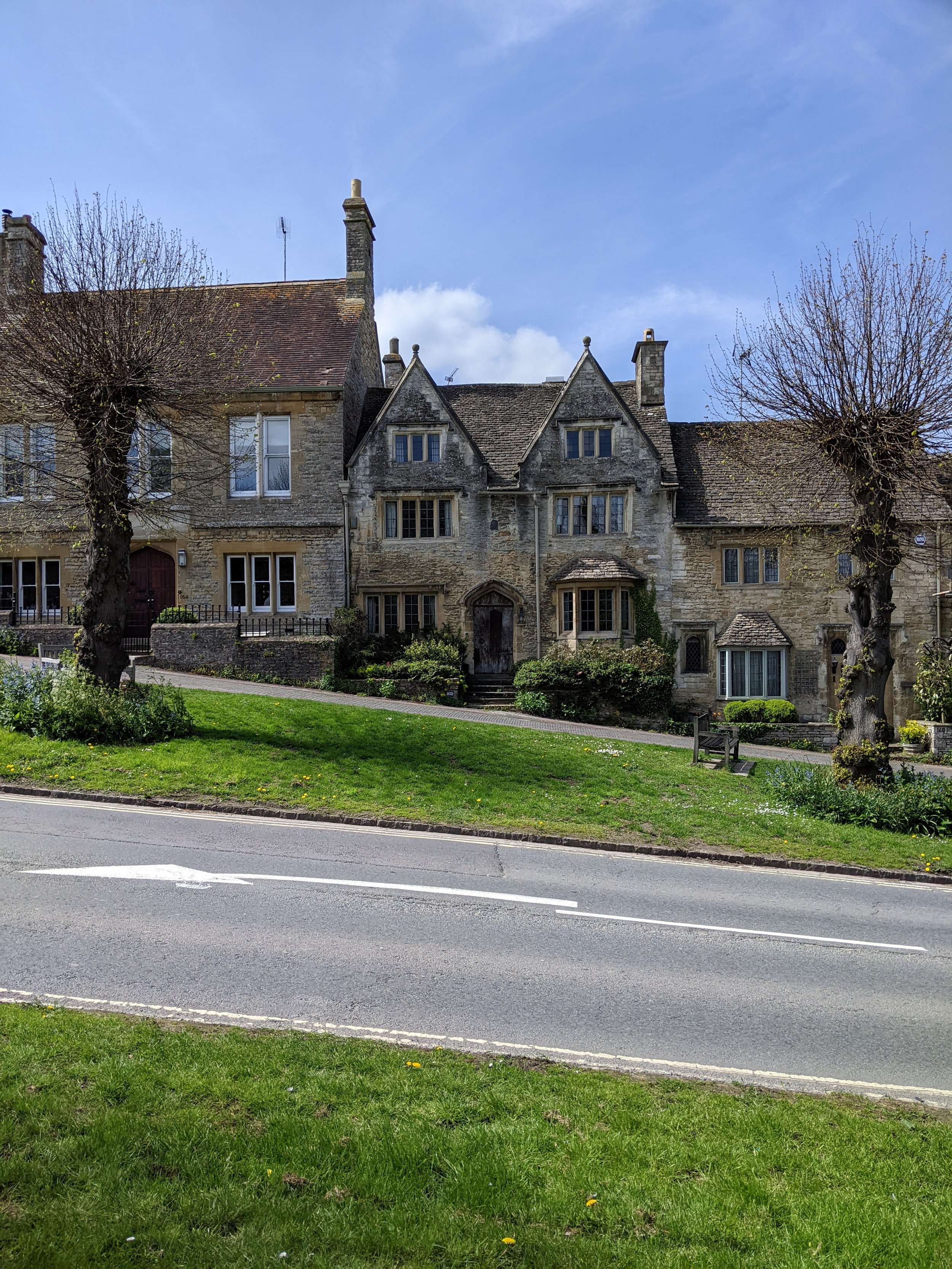 Burford, Cotswolds. Stone houses with multiple gables, tall chimneys, and leafless trees in front, situated along a street with a grassy area and a pavement.