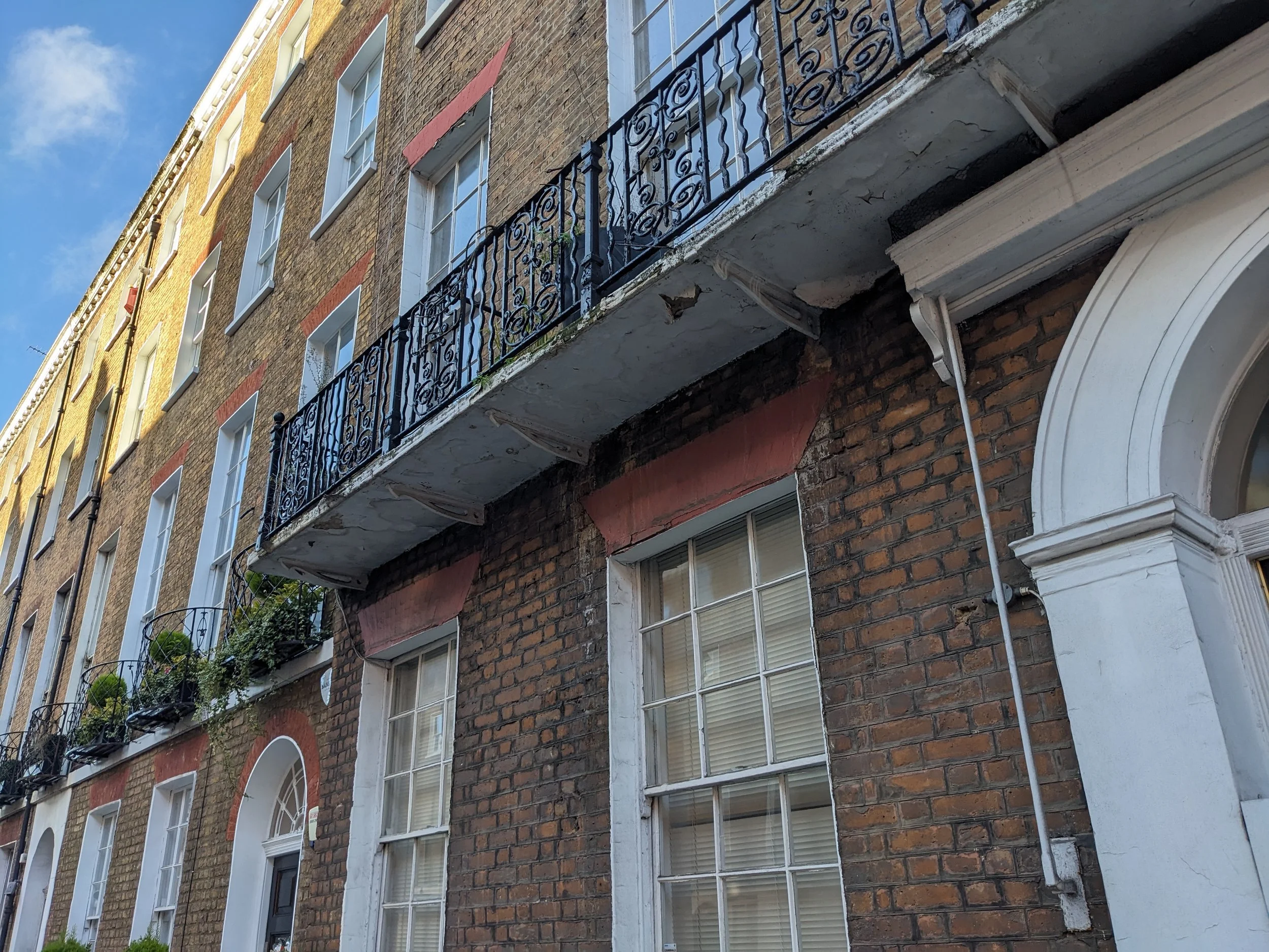 Close-up of Regency London terraced townhouses with balcony at piano nobile. Timber sash windows under brick voissiors. Under a blue sky with some clouds.