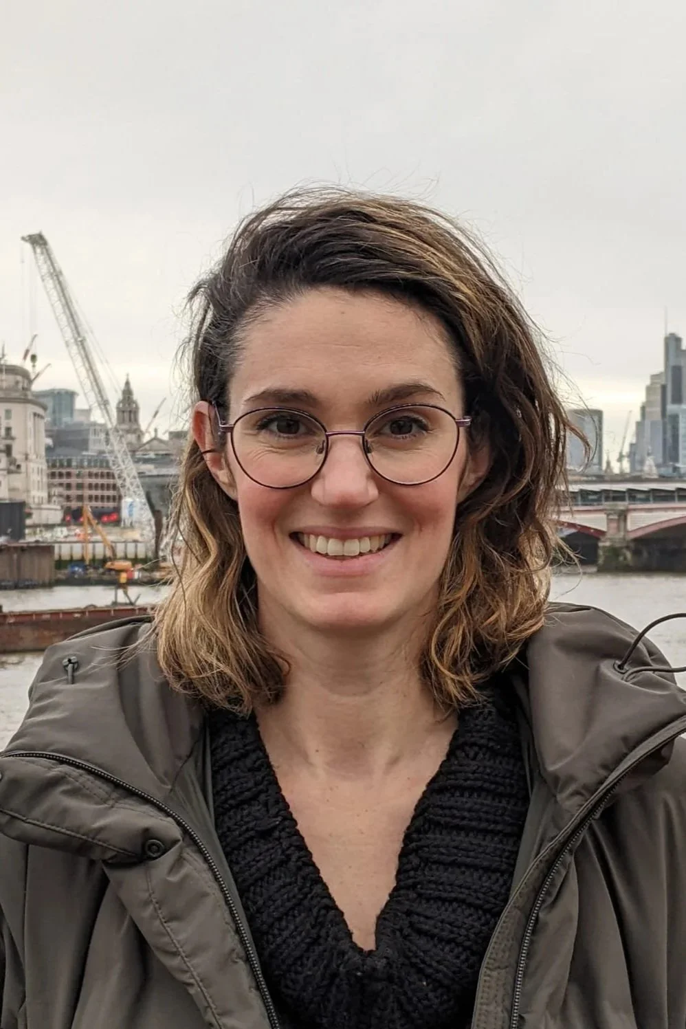 Founder of Pelta Heritage. A woman with shoulder-length wavy brown hair, wearing glasses and a grey jacket, smiling outdoors with a city skyline and river in the background.