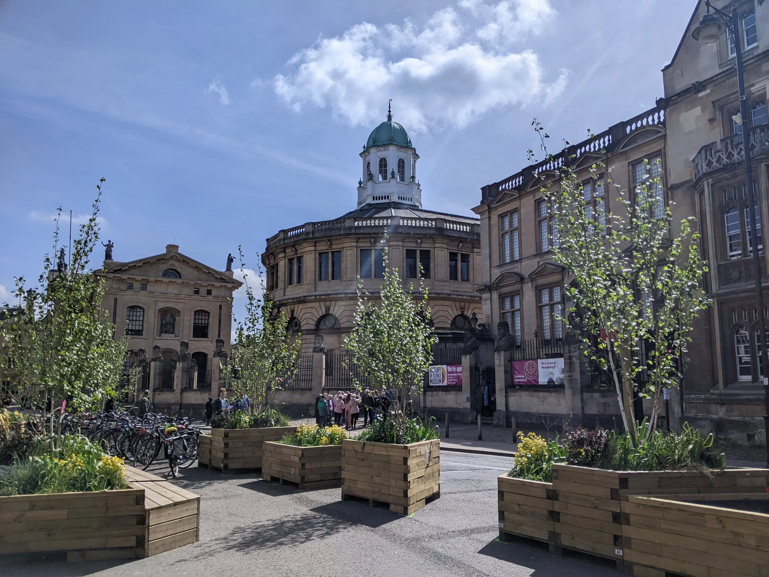 Urban park with planters, trees, and historical buildings with a green-domed tower in the background, on a sunny day.