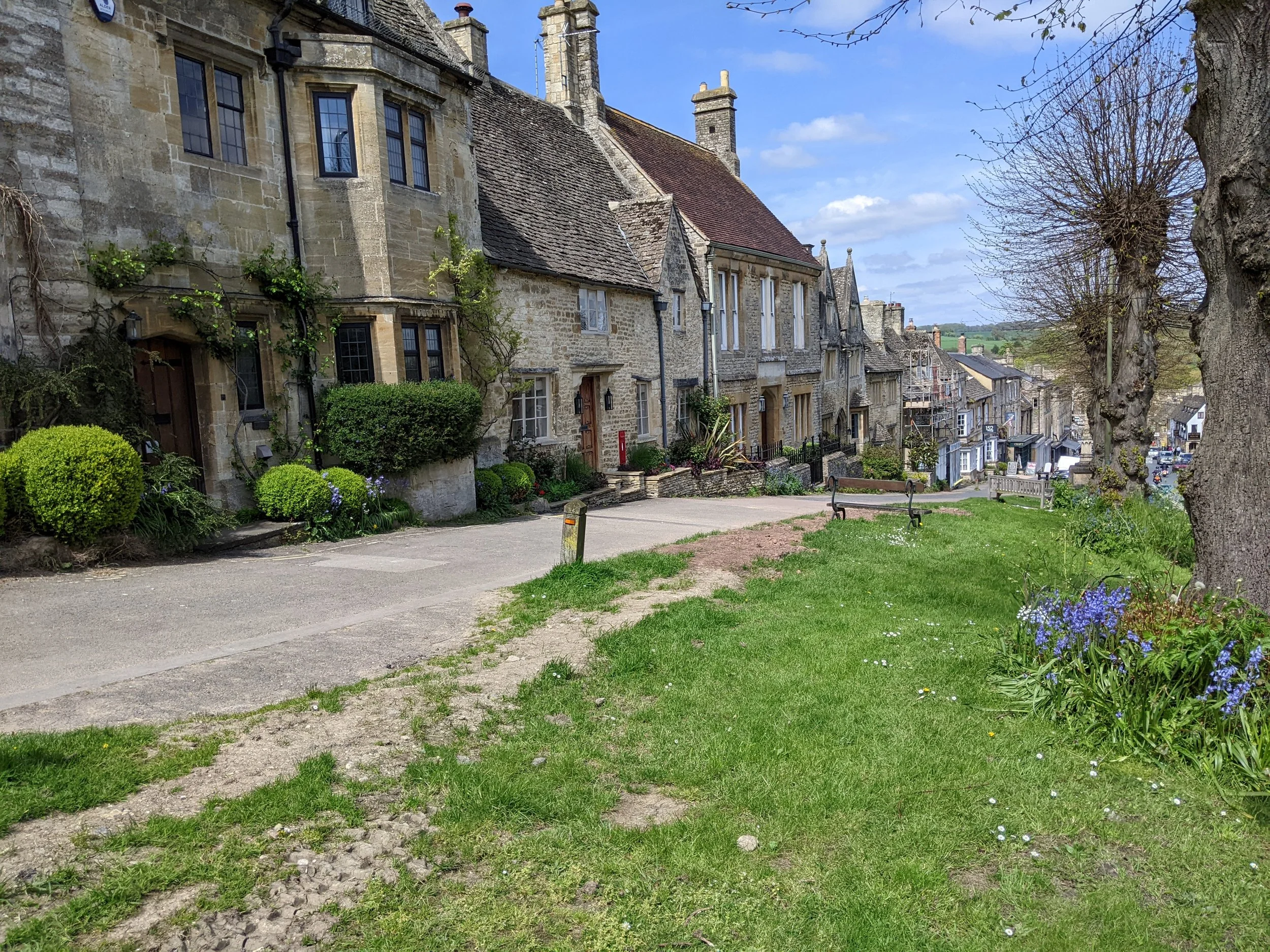View of historic stone buildings with tiled roofs, small trees, and flowers along a street under a blue sky with some clouds.