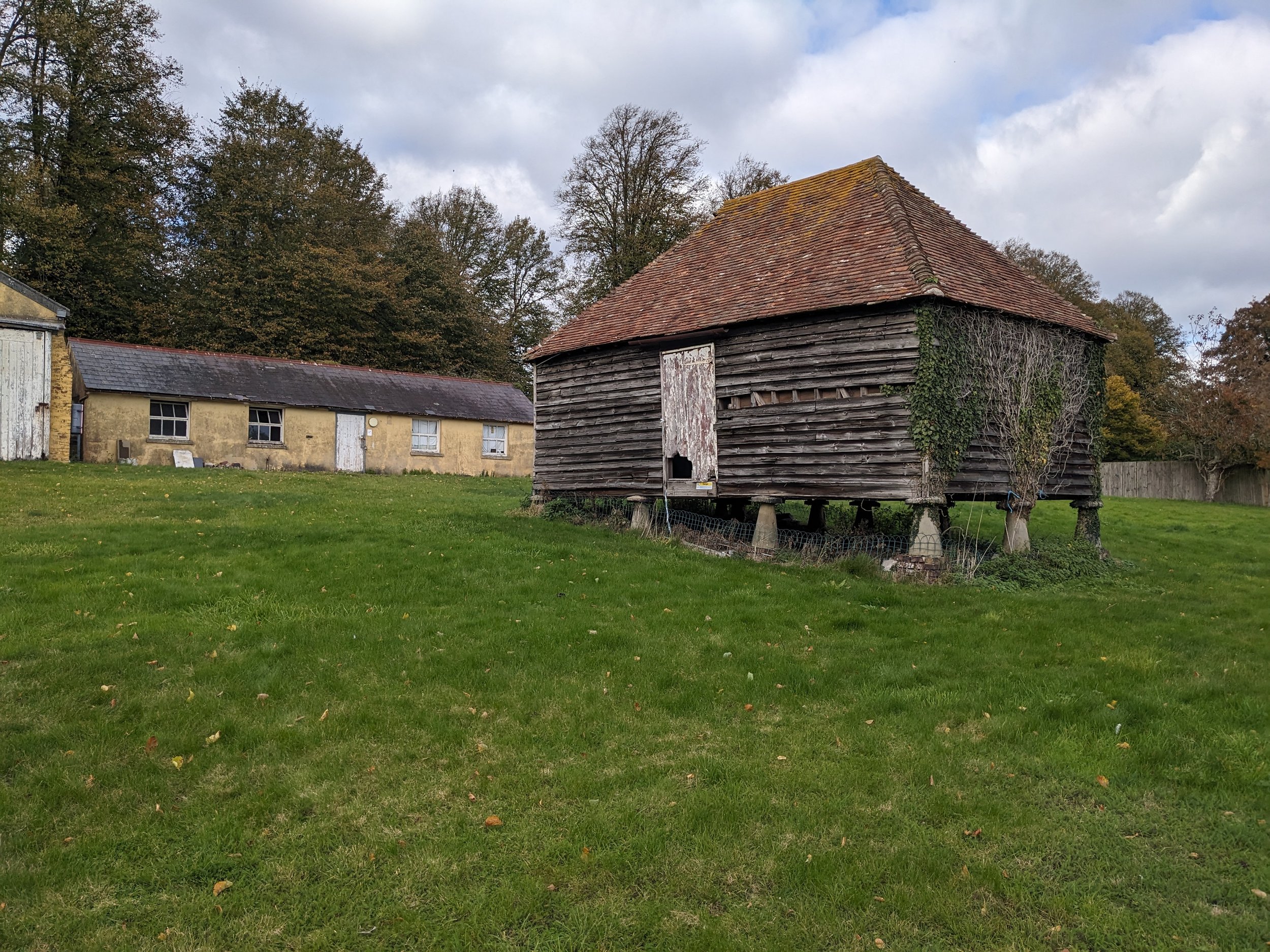 An old wooden barn with a shingled roof, surrounded by trees with fall foliage, and a yellow house with white windows in the background, under a cloudy sky.