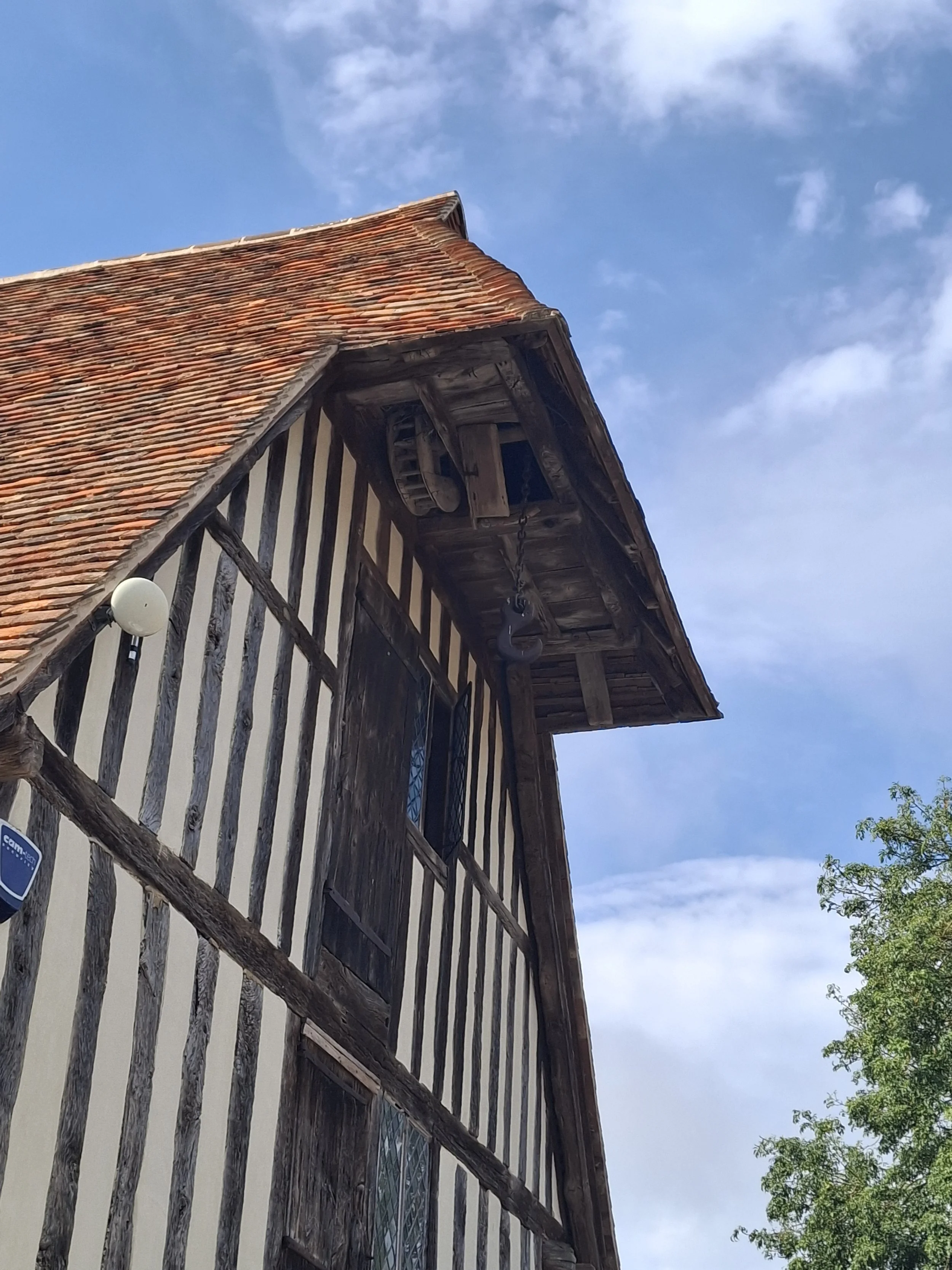 An old traditional half-timbered building with a steep red tile roof, wooden beams on the facade, and a chain mechanism hanging from the upper balcony, under a partly cloudy blue sky.