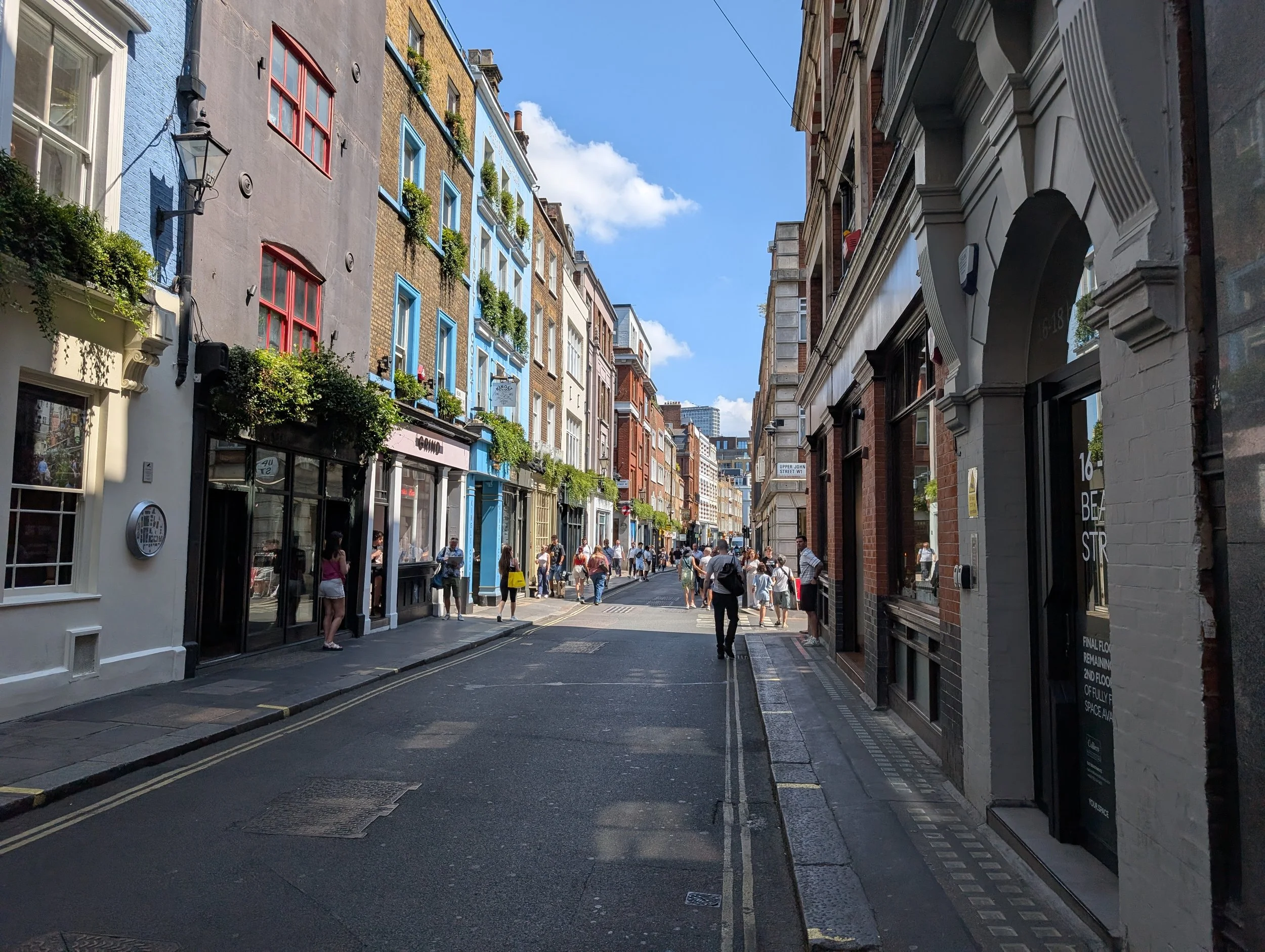 A busy Soho, London, street scene with colorful buildings on both sides and people walking along the pavement under a blue sky with some clouds.