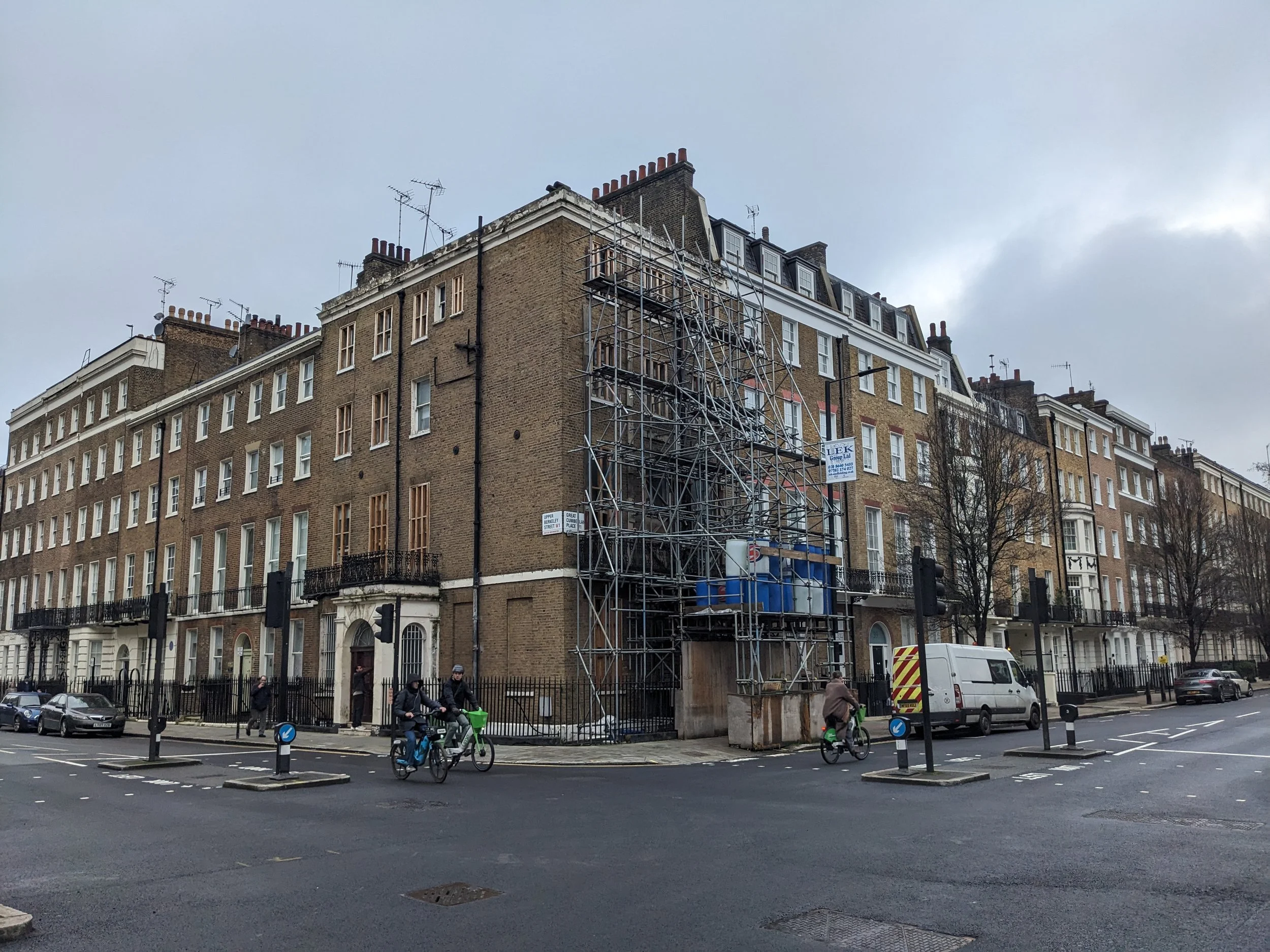 Street view with row of multi-story brick apartment buildings, some scaffolding on a building, cars parked along the street, people riding bicycles, and a cloudy sky overhead.