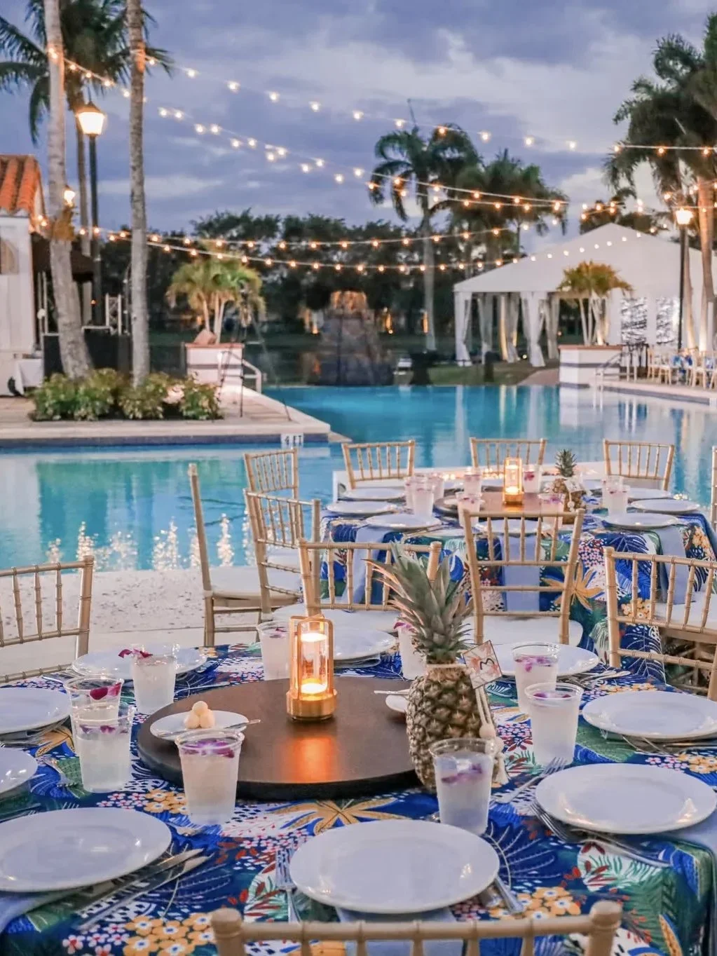 Outdoor poolside dining setup with tropical-themed tablecloths, pineapple centerpiece, and string lights at dusk.