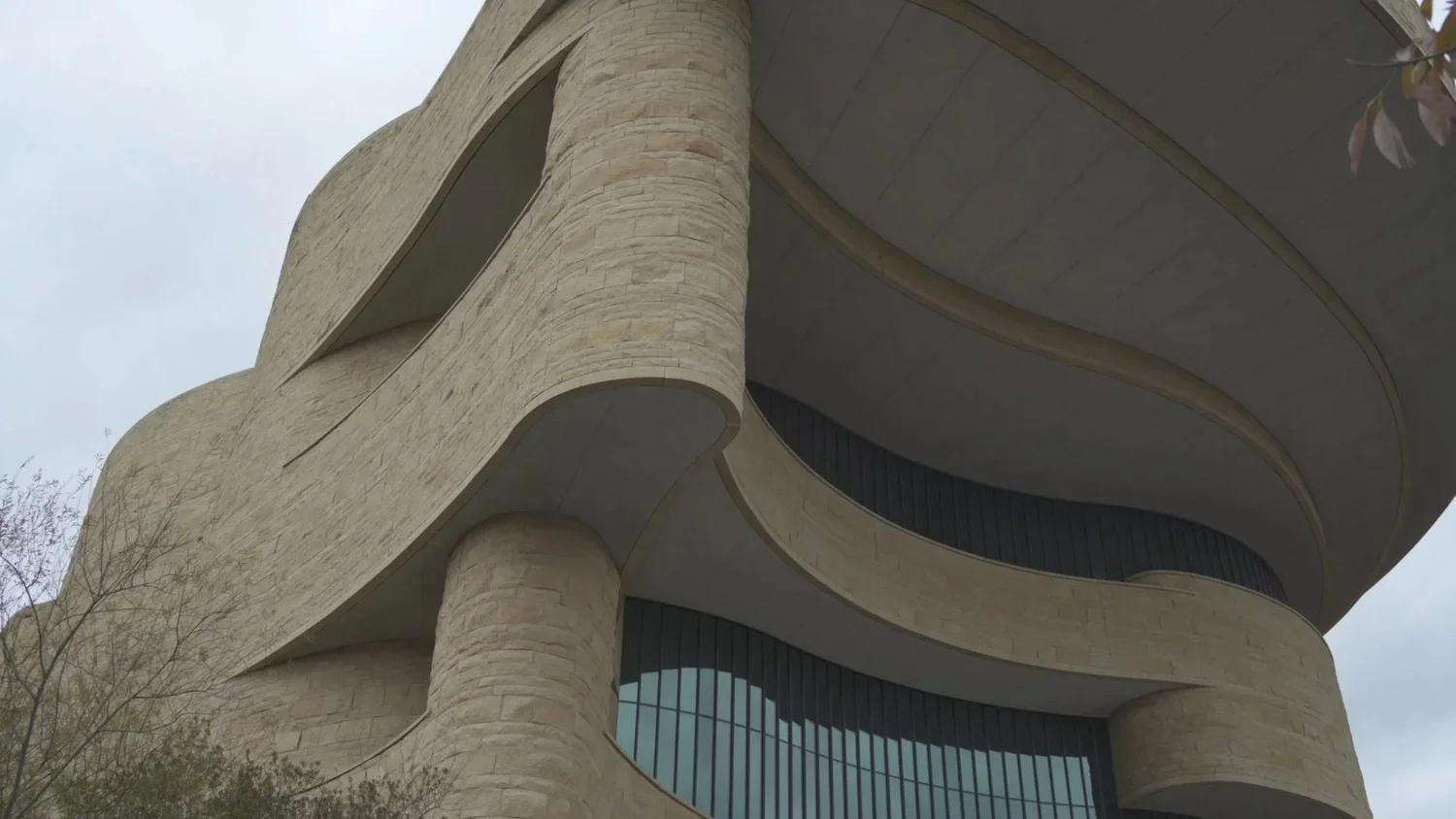 Close-up of a modern building with curved, beige stone walls and large glass windows, viewed from below.