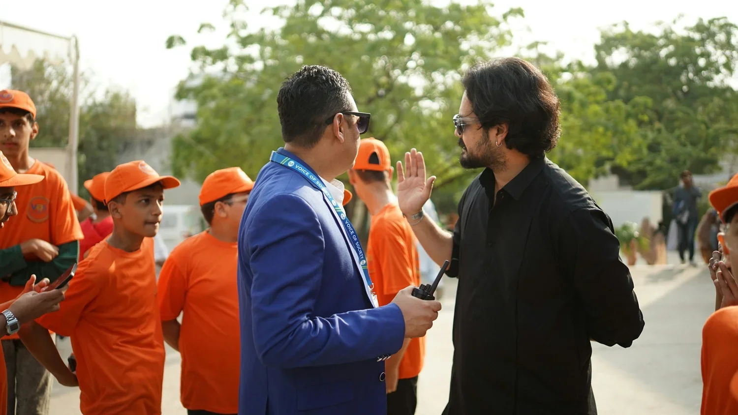 Two men are having a conversation outdoors; one in a blue suit holding a walkie-talkie, and the other in black, raising his hand in a gesture, with a group of children in orange uniforms and caps in the background.