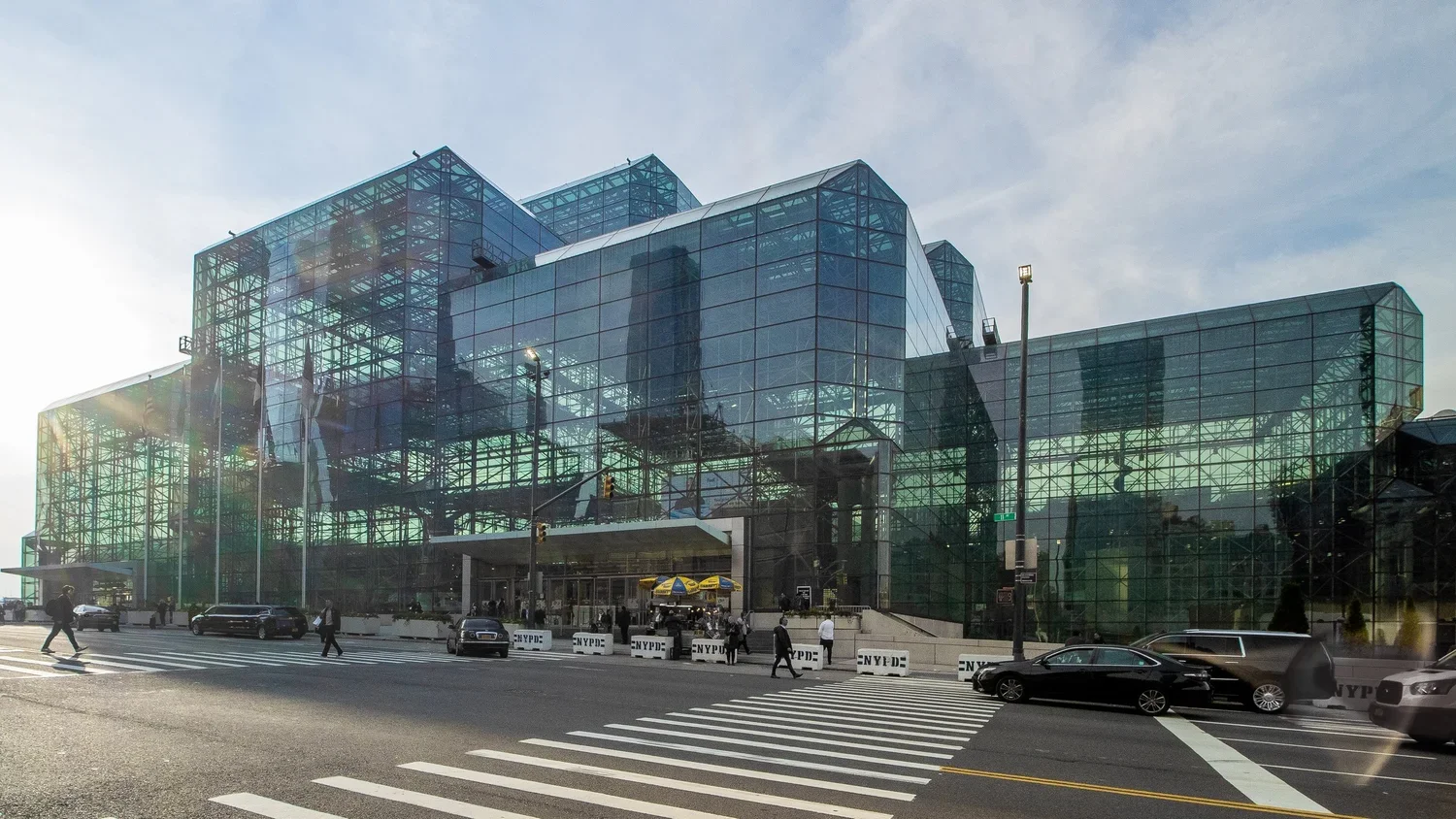 Modern glass building, possibly a museum or office, with pedestrians and cars crossing the street in front.