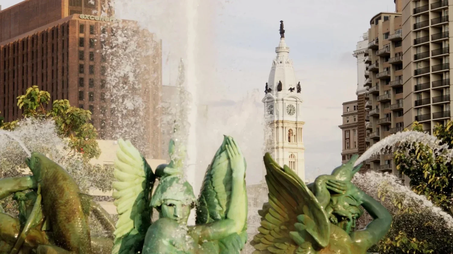 Fountain with sculptures of winged figures spraying water, with city buildings and a clock tower in the background.