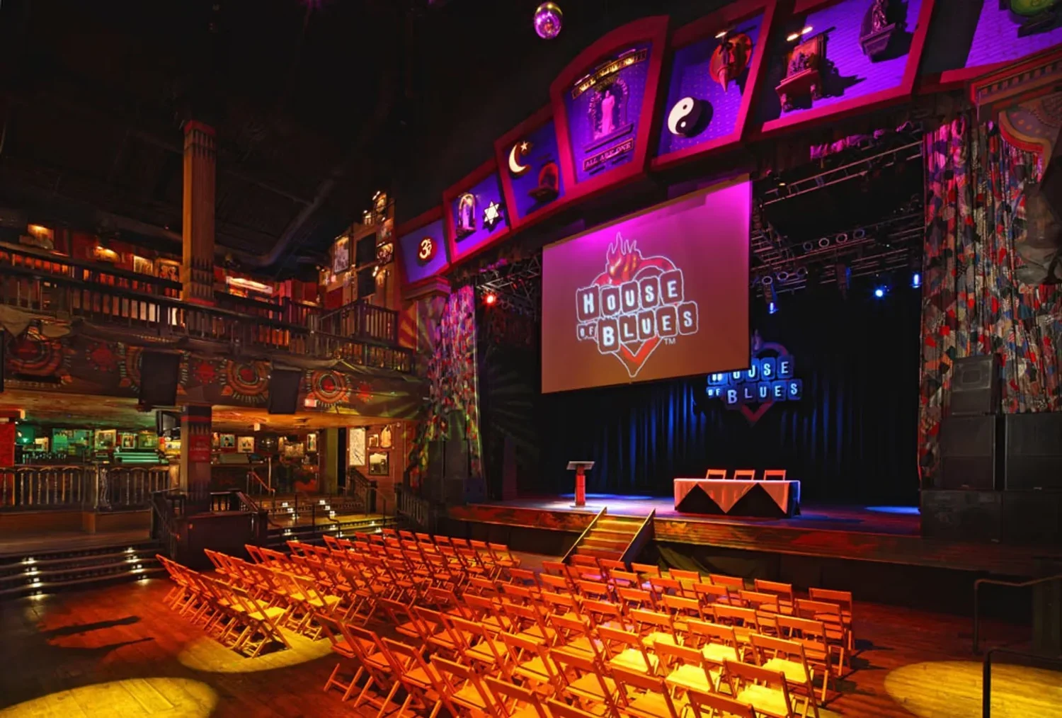 Empty stage with a backdrop displaying the 'House of Blues' logo, surrounded by rows of orange chairs in front, in a colorful, dimly lit concert venue with balcony seating.