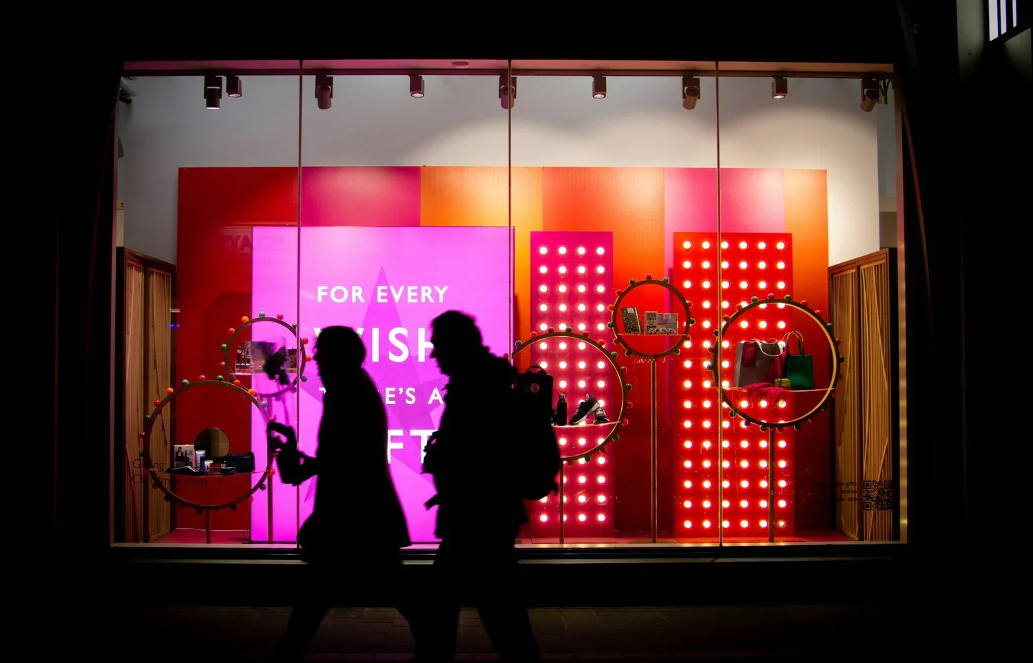 Silhouettes of two people walking past a brightly lit store display window with colorful pink and red decor, illuminated circles, and bags on display.