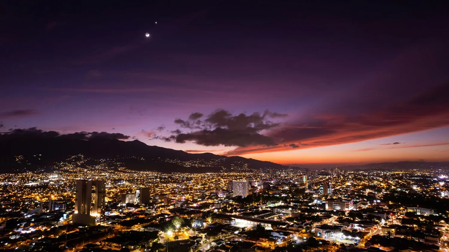 Nighttime cityscape with illuminated buildings, a mountain in the background, and a colorful sunset sky with a crescent moon and visible planets