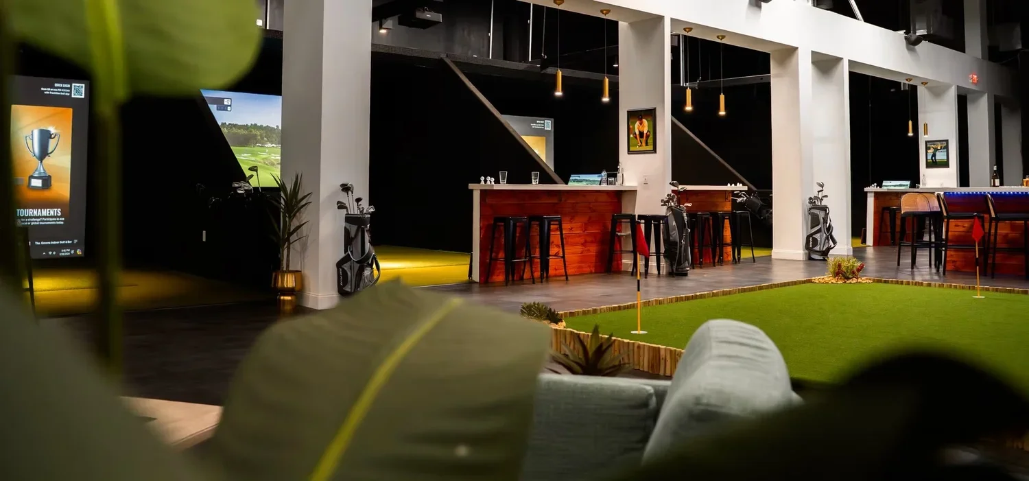 Indoor golf simulation lounge with a mini putting green, golf bags, and a bar area with stools, illuminated by hanging pendant lights.