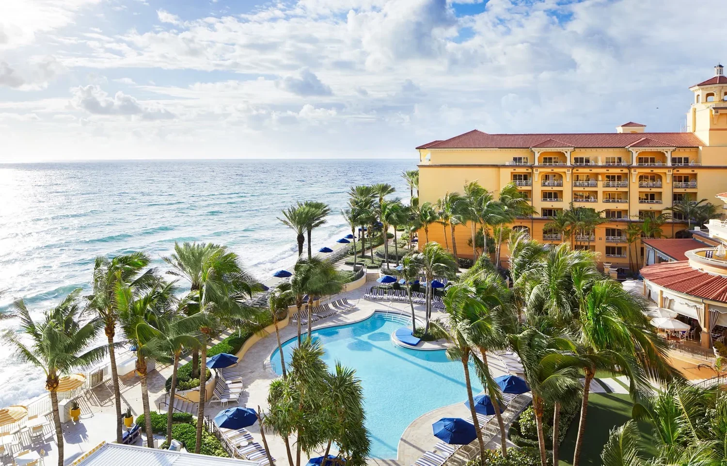 Aerial view of a resort with a swimming pool surrounded by palm trees, lounge chairs, and umbrellas, with an ocean backdrop and a large yellow hotel building.