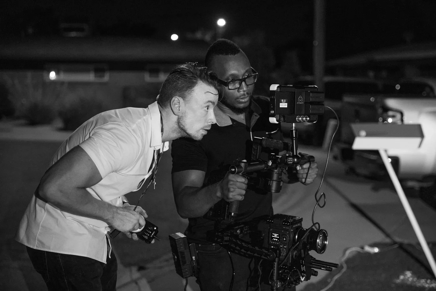 Two filmmakers review footage or shots on a night outdoor set with a camera rig, one holding a remote control, in black and white.