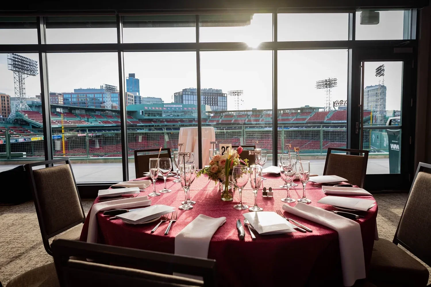 Round banquet table with red tablecloth, white napkins, and glassware set for dining, overlooking a baseball stadium through large glass windows.