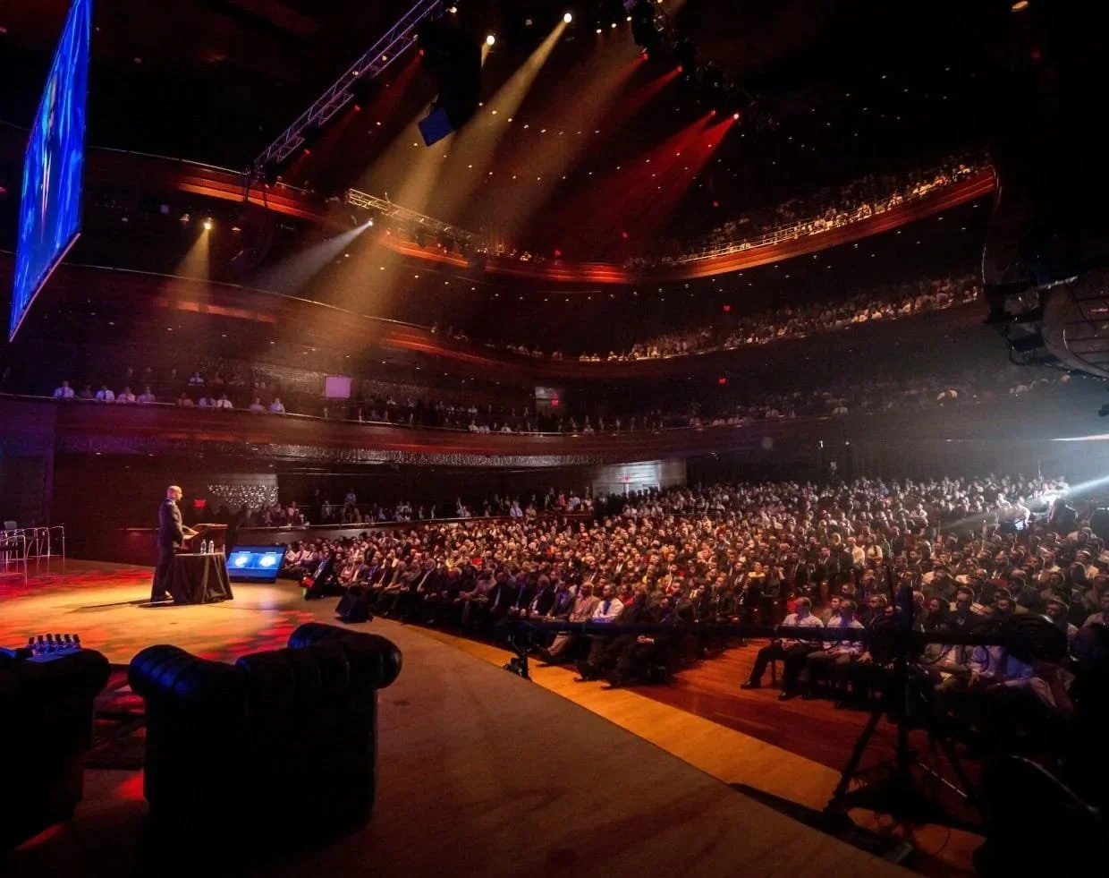Large auditorium filled with standing and seated audience, stage with a speaker at a podium, colorful stage lighting, multiple levels of balcony seating, and a large screen on the left.