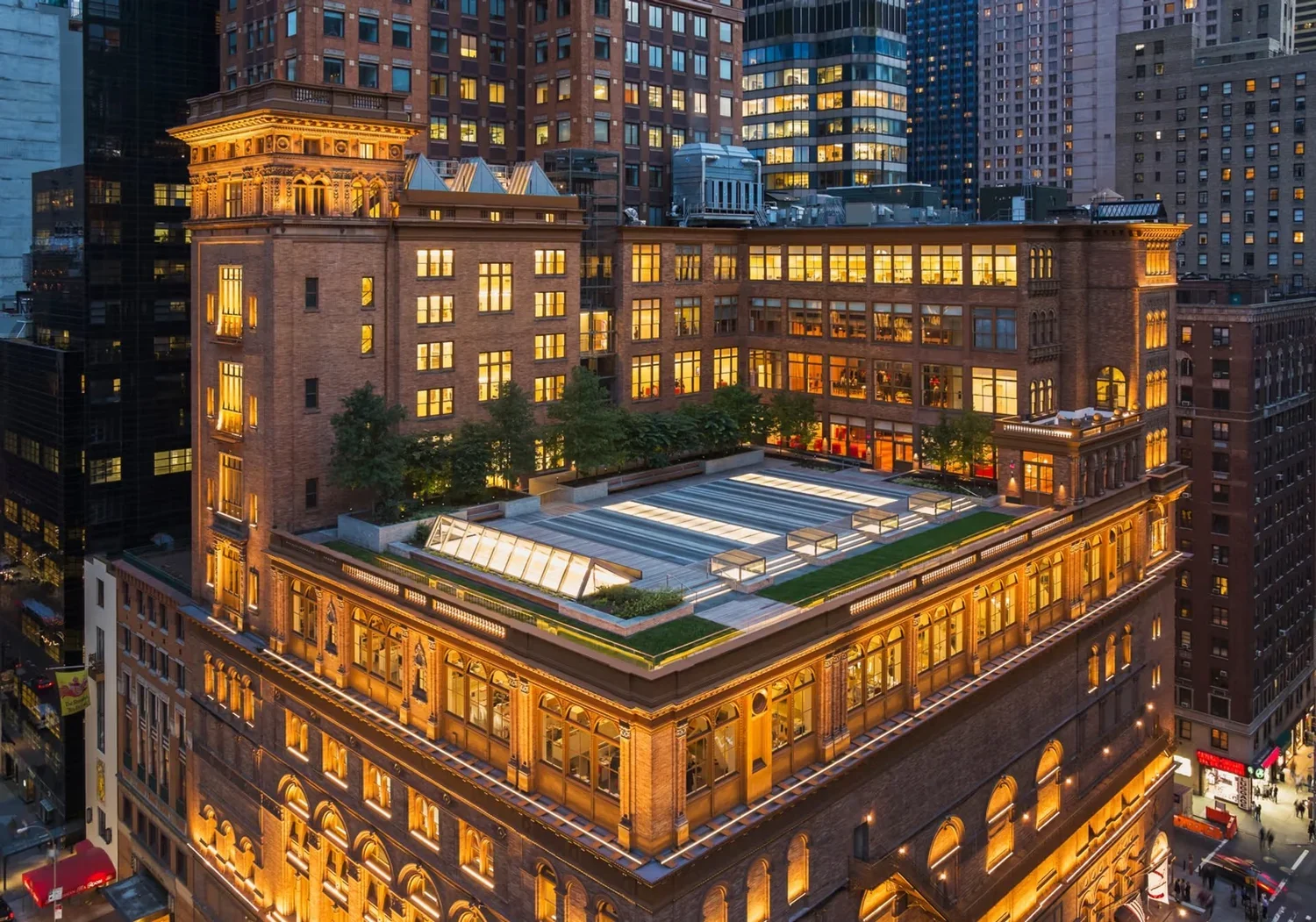 Aerial view of a city skyscraper at dusk with illuminated windows, rooftop garden, and surrounding tall buildings.
