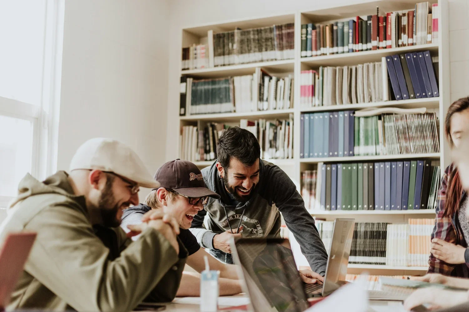 Four young men are laughing and working together at a table with laptops in a library or study room.