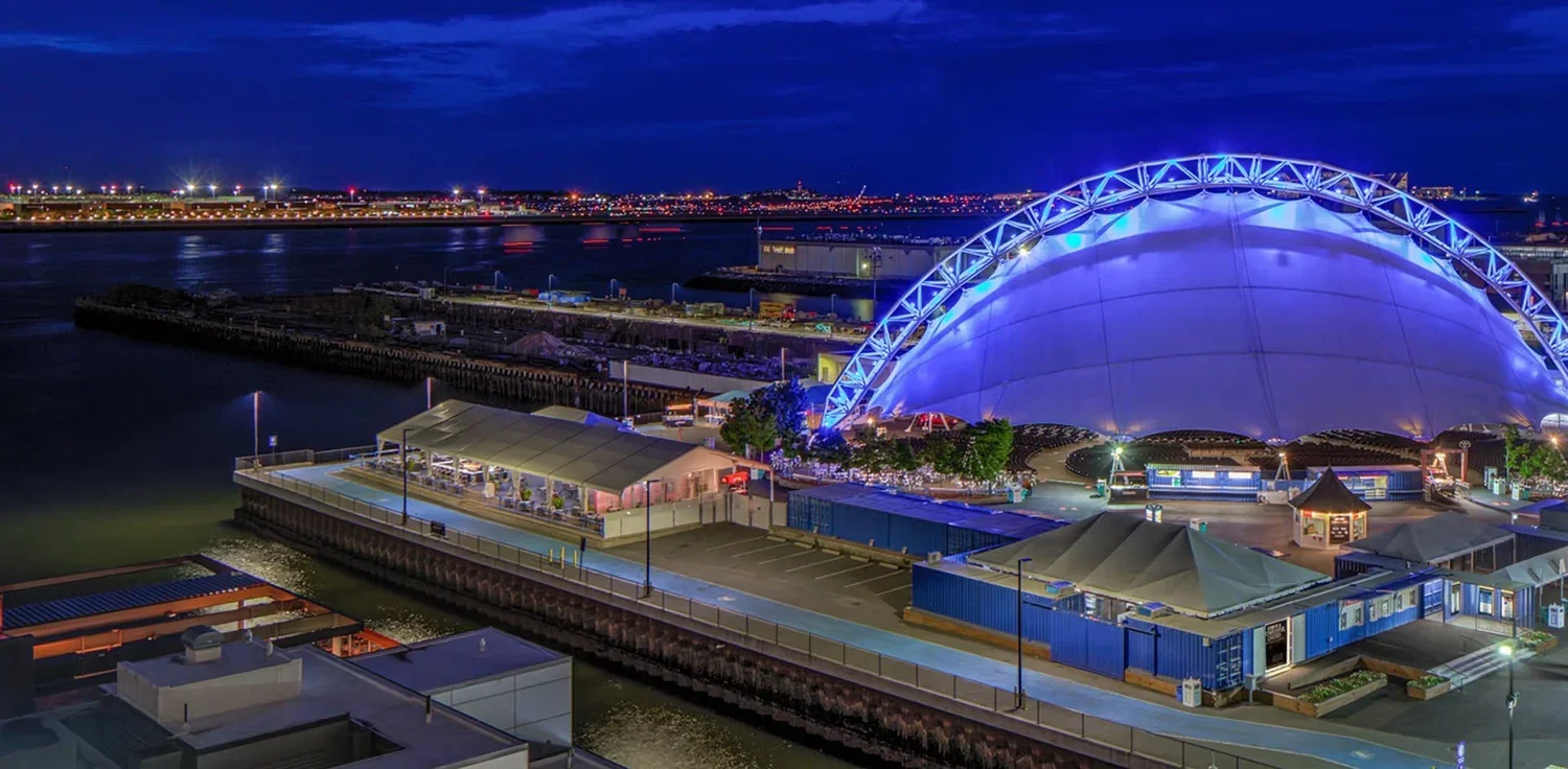 Night view of an illuminated waterfront entertainment venue with a large dome structure lit in blue, surrounded by smaller buildings and parking areas.