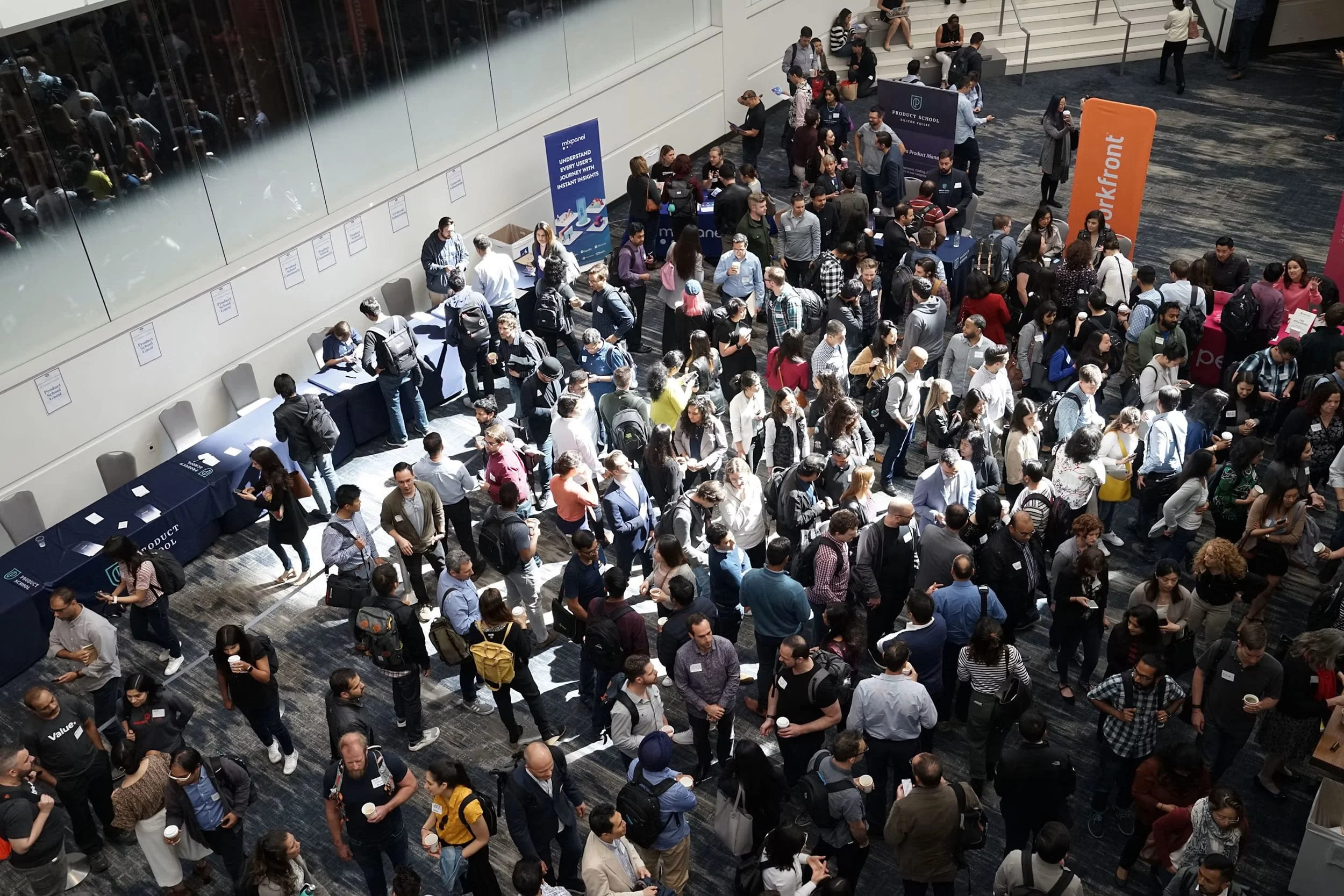 A crowded conference or event hall with numerous people walking and standing in lines. Several booths and banners are set up, including one with the text 'WORKfront' and others with branding and informational materials. People are holding coffee cups, engaging in conversations, and using their phones.