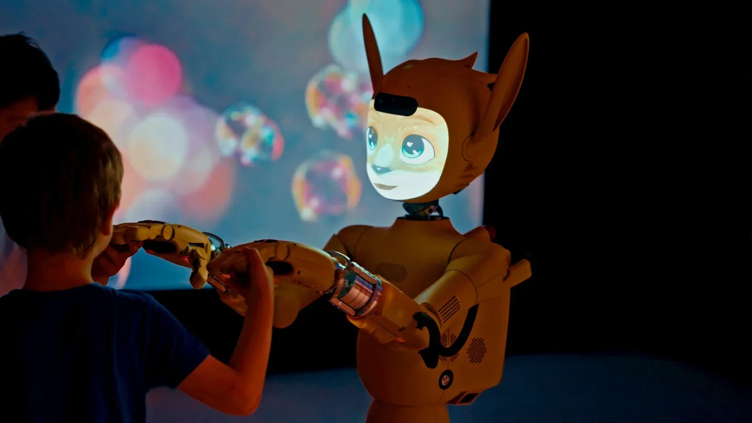 A robot with a humanoid face and animal-like ears extends a hand to a child in a dark room with a colorful, bokeh background projected on the wall.