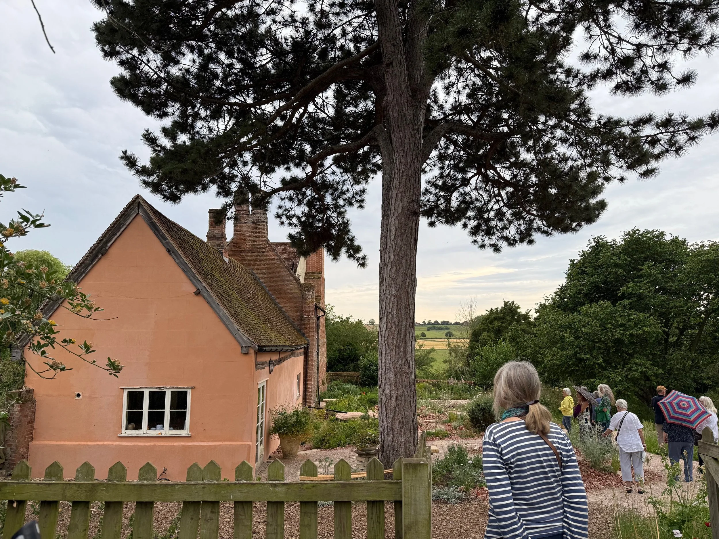 People gathered outside a peach-colored house surrounded by trees, with some holding umbrellas, under an overcast sky.