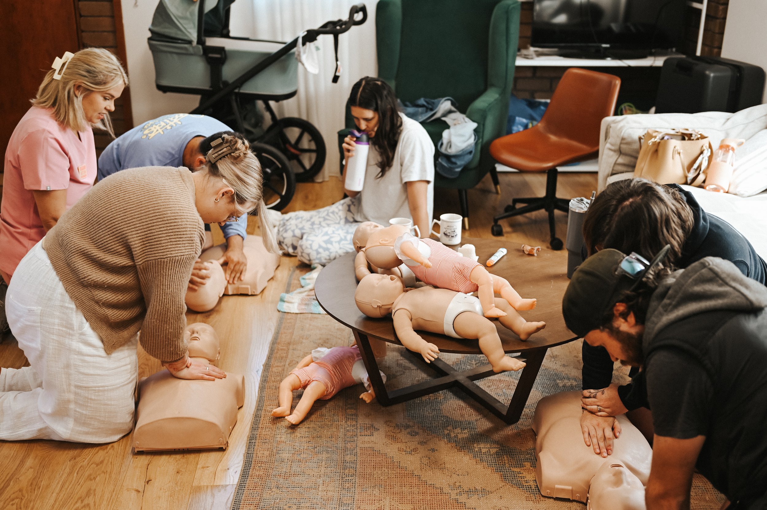 People participating in CPR training with mannequins on a wooden floor, practicing chest compressions and rescue techniques in a room with chairs, a table, and medical supplies.