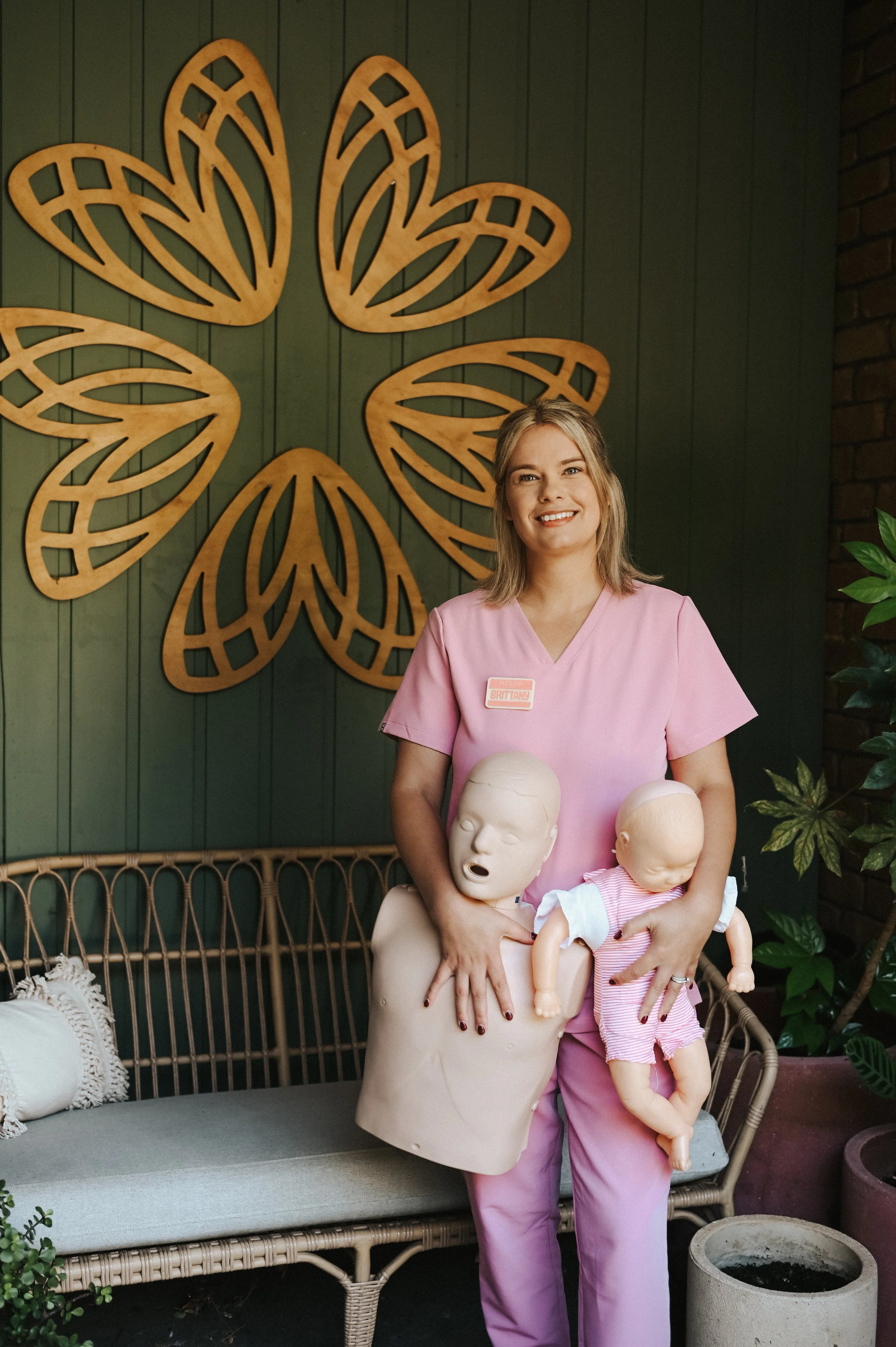 A woman dressed in pink scrubs holding two mannequins of babies, one with its mouth open and another with closed eyes, standing in a room with a green wall decorated with wooden moth art and surrounded by plants at the mums of the hills hub