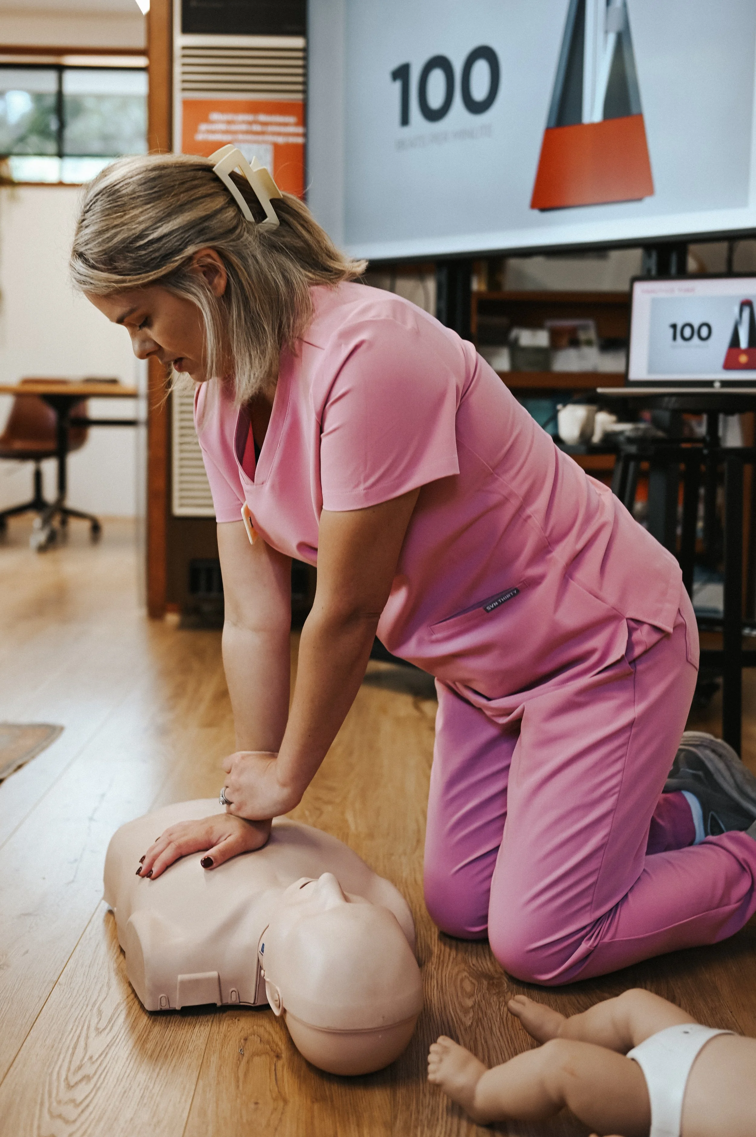 A paediatric nurse in pink scrubs practicing baby and child CPR on a medical dummy during a training session for a mothers group at the mums of the hills hub